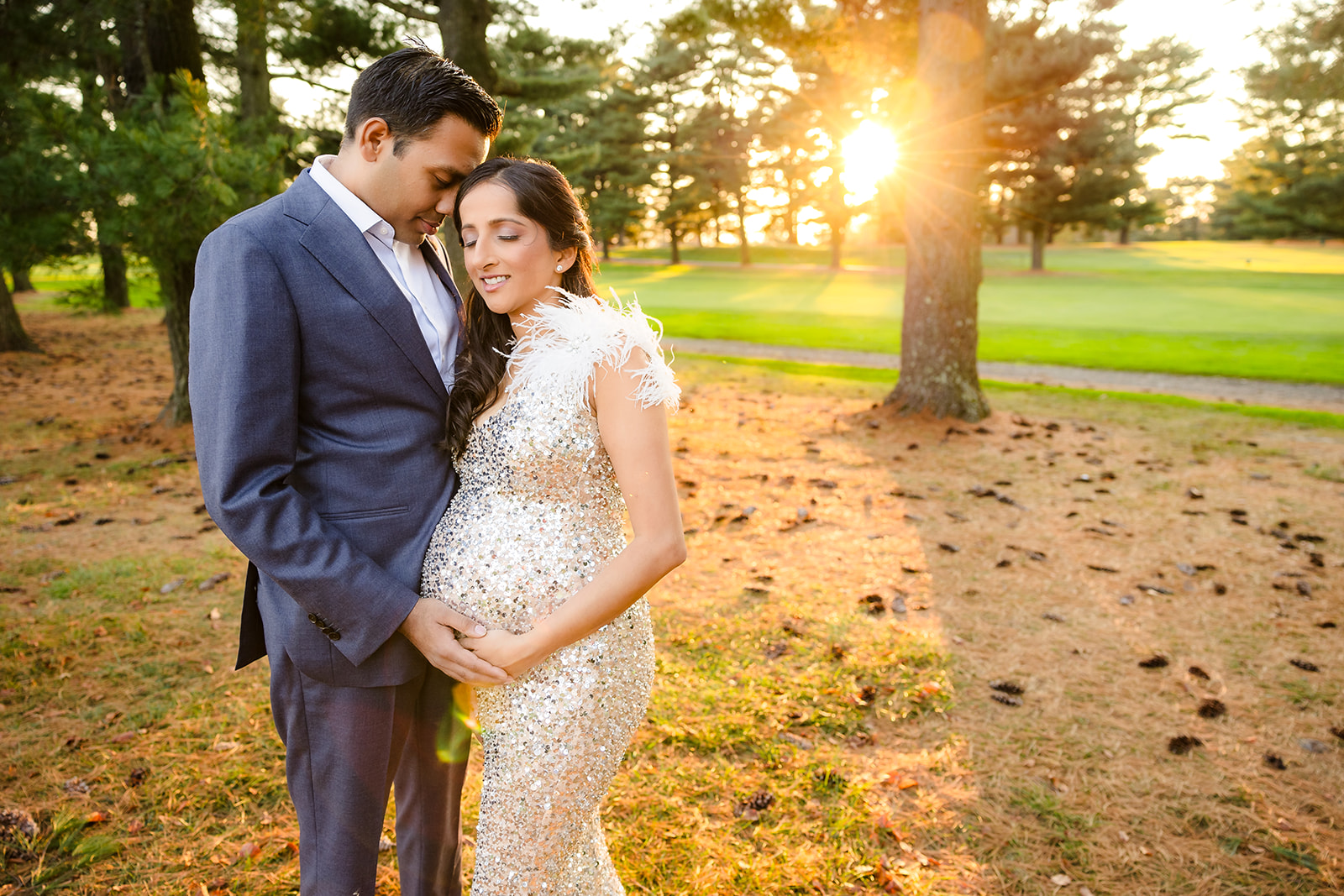 Couple embracing and holding pregnant belly with the sun in the back and their eyes closed.